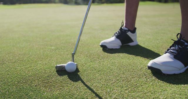 Close-up of golfer's feet and putter poised to hit golf ball on manicured green. Ideal for sports articles, golfing guides, athletic gear advertisements, or promotional materials for golfing events.