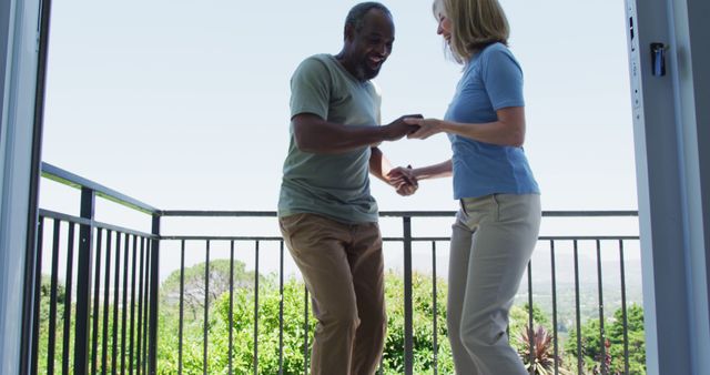 A senior couple is dancing joyfully on a balcony with a scenic view of greenery and mountains. This lively and heartwarming image can be used in advertisements promoting active lifestyles, retirement communities. It is also suitable for materials related to health, wellness, and senior activities.