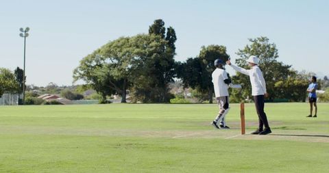 Cricket umpire raising arm while batsman stands at stumps on sunny suburban green outfield