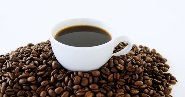 A white cup filled with coffee sits atop a mound of roasted coffee beans, with copy space. The contrast between the dark brew and the white background emphasizes the rich color of the coffee.