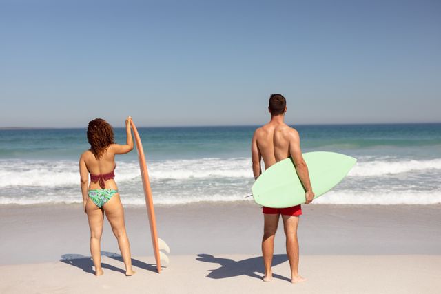 Diverse couple standing on beach holding surfboards, looking at ocean waves. Ideal for promoting summer vacations, surfing activities, beach lifestyle, and outdoor fitness.