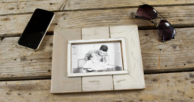 Framed family portrait sits on an aged wooden table, complemented by a smartphone and sunglasses nearby. The rustic wood frame holds a black and white photo of an adult and a child sharing a moment. Ideal for use in themes related to family, nostalgia, or personal moments. Suitable for articles, blogs, or advertisements that emphasize family bonds, simplicity, rustic aesthetics, or lifestyle.