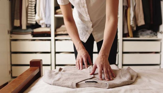 Woman folding sweater in minimalist wardrobe organizing cozy knit on bed for decluttering