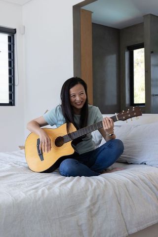 Mature asian woman playing acoustic guitar on bed in minimalist bedroom, relaxing hobby