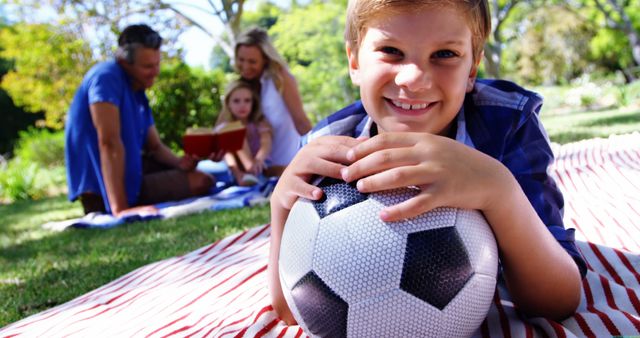Young boy holding soccer ball while smiling and enjoying family picnic in park. Background includes family members sitting on blanket, reading a book. Great for use in advertisements promoting family activities, outdoor fun, sports, and healthy lifestyle. Ideal for articles or websites about parenting, leisure, and recreation.