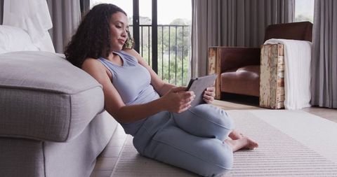 Woman relaxing on rug leaning against sofa using tablet and reading messages in minimalist home