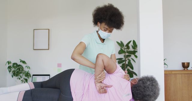A physical therapist is aiding a senior woman with a spinal exercise in a bright, indoor setting. Both individuals are wearing face masks, emphasizing health and safety precautions. This can be used to illustrate themes of rehabilitation, healthcare, and wellness, or to promote senior care services, physical therapy programs, and active aging initiatives.