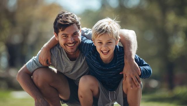 Smiling father and son crouching together in sunlit park sharing joyful outdoor bond