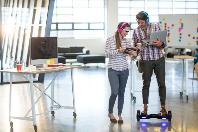 Two colleagues in a modern office environment are using digital devices. One is holding a tablet while the other is balancing on a hoverboard with a laptop. Both are wearing headphones, indicating a tech-savvy and innovative workspace. This image is ideal for illustrating concepts of modern work culture, technology integration, and collaborative teamwork in a startup or creative office setting.