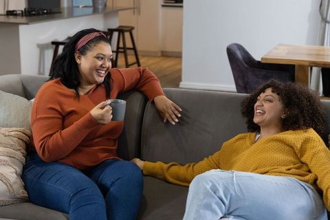 Laughing diverse female friends relaxing on sofa with coffee, cozy home conversation