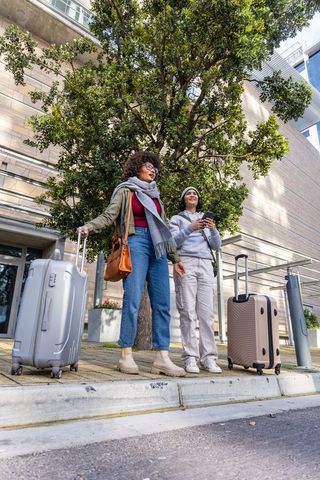 Female friends waiting curbside with luggage outside modern building holding smartphone