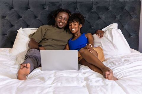 African american couple relaxing in bed using laptop together smiling cozy modern bedroom