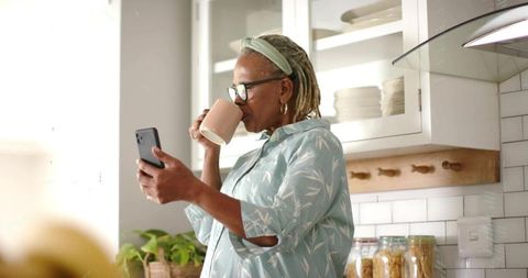 Senior woman enjoying morning coffee while checking smartphone in sunlit kitchen
