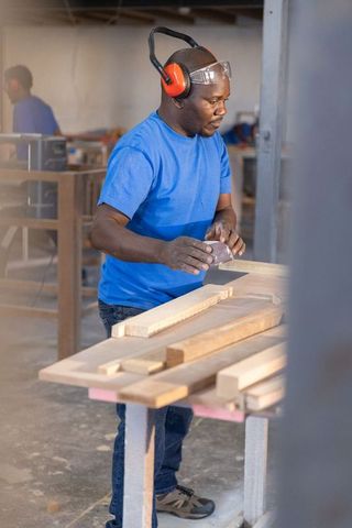 Carpentry workshop worker sanding wood plank wearing ear protection and safety goggles
