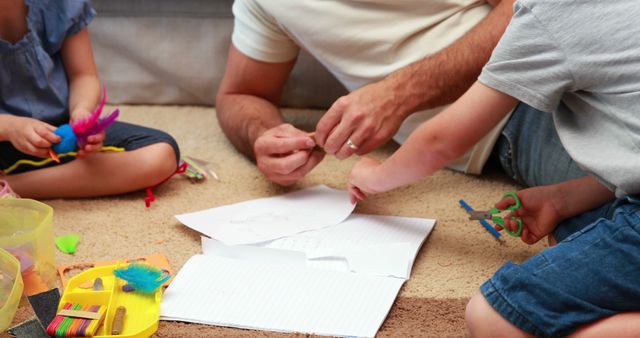 Father and children participating in creative arts and crafts activities at home, using paper, markers, scissors, and colorful feathers. This family scene highlights creativity, educational play, and family bonding time. Perfect for use in articles or ads focusing on parenting, home education, childhood development, or family activities.