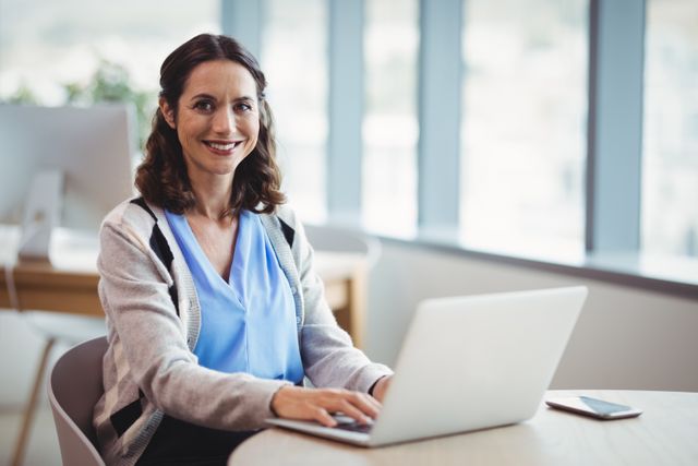 Smiling businesswoman using laptop while sitting at desk in a modern office. Great for promoting workplace productivity, corporate culture, technology in business, and professional settings.