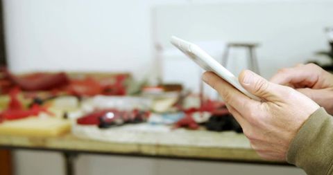 Hands Holding Smartphone at Workbench Tapping Screen with Tools and Red Parts Nearby