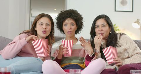 Three women watching movie on couch eating popcorn and reacting with surprise