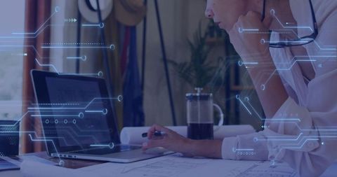 Mature woman reviewing laptop and blueprint plans with digital HUD overlay in home office
