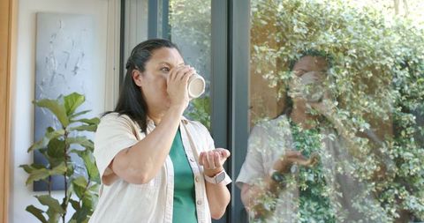 Mature african american woman sipping coffee by glass door with smartwatch and leafy reflection