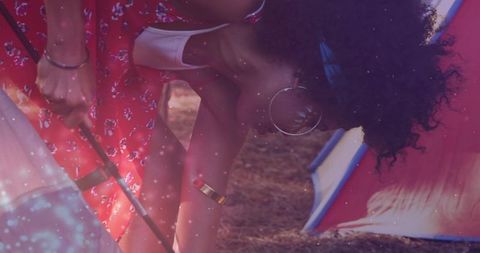 Curly-haired woman adjusting tent pole while friend in red dress holds support for camping