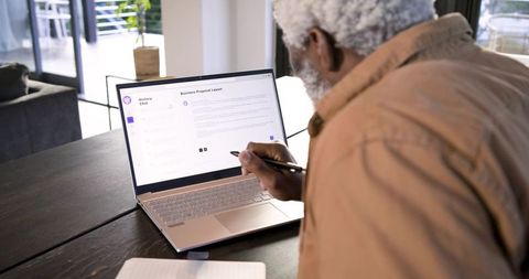 Senior man writing in notebook using laptop at home office
