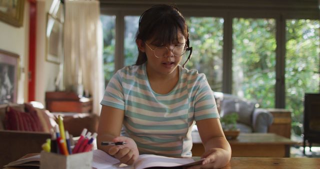 Teenager wearing glasses and headset while studying with an open book at home. Ideal for educational content, remote learning advertisements, academic websites, and online tutoring services.