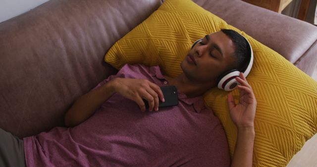 Young man lying on a brown couch, wearing headphones and holding a smartphone against a yellow pillow. Perfect for themes like relaxation, leisure, technology, music streaming, and personal comfort.