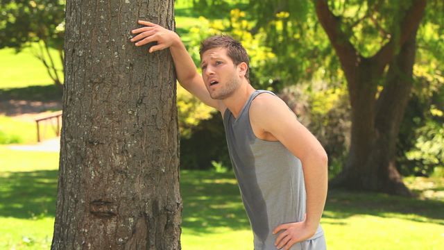 A man wearing sports attire leans against a tree, looking exhausted after a jog in a scenic park filled with lush greenery. His attire is dark athletic wear appropriate for physical activity. This scene exemplifies a healthy lifestyle and could be useful in fitness programs, health blogs, or outdoor activities promotional material showcasing the benefits of daily exercise in natural settings.