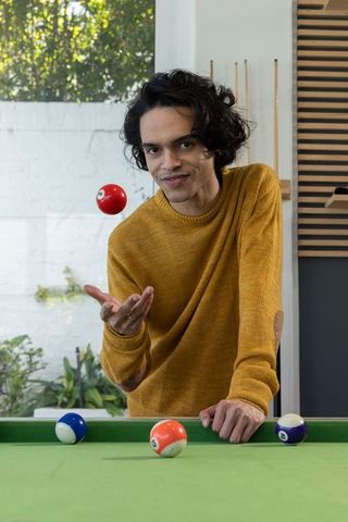Young man tossing red billiard ball over pool table in modern game room with cue rack