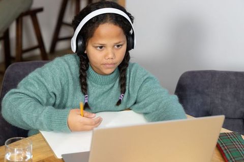 Preteen girl studying remotely wearing headphones using laptop and notebook