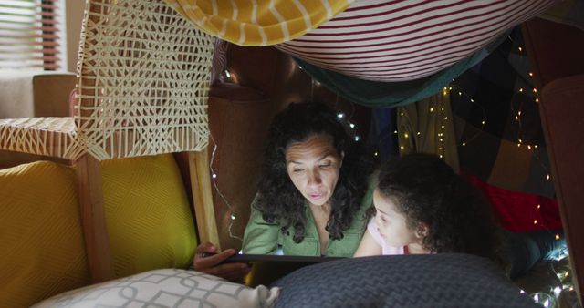 Mother and daughter enjoy a story together inside a cozy blanket fort adorned with pillows and fairy lights. Perfect for themes of family bonding, indoor activities, parent-child relationships, and imaginative play. Great for use in advertisements, parenting blogs or articles, and marketing materials focused on family-friendly content.