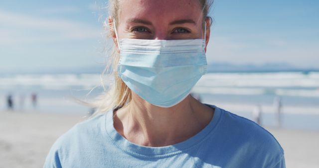 Young woman wearing a face mask, smiling at camera while standing on a beach during a sunny day with the ocean in the background. Perfect for themes on outdoor safety, pandemic, healthy lifestyle, and travel. Useful for websites, blogs, advertisements, and public health campaigns.