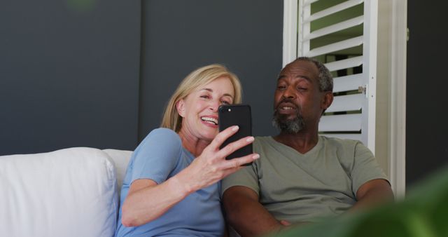 This image depicts a happy couple sitting on a white couch, sharing a moment while looking at a smartphone. It is perfect for illustrating technology use in daily life, representing a diverse relationship, or promoting products and services related to digital connectivity, leisure time, and modern living rooms.