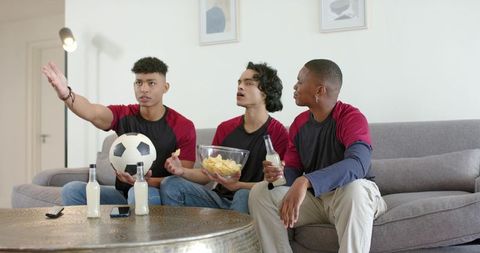 Young friends watching soccer match on couch holding soccer ball chips drinks cheering