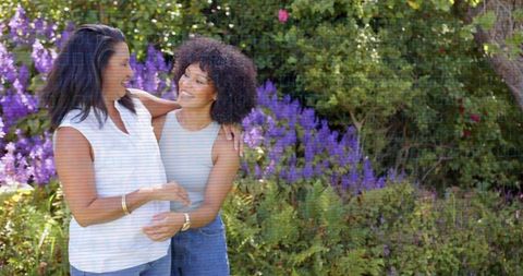 Smiling mother and daughter hugging in sunlit garden with purple blooms and greenery