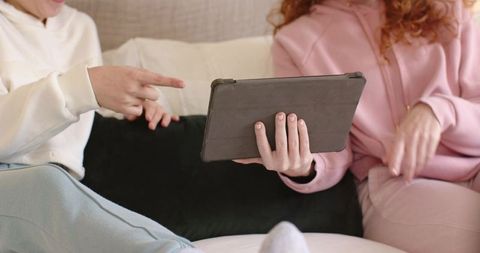 Diverse female friends sharing tablet content on couch while wearing hoodies and cozy socks
