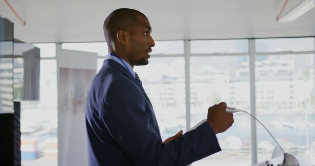 Side view close up of a young African American businessman wearing a suit and tie standing at a lectern holding a tablet, gesturing with his fist and addressing the audience at a business conference