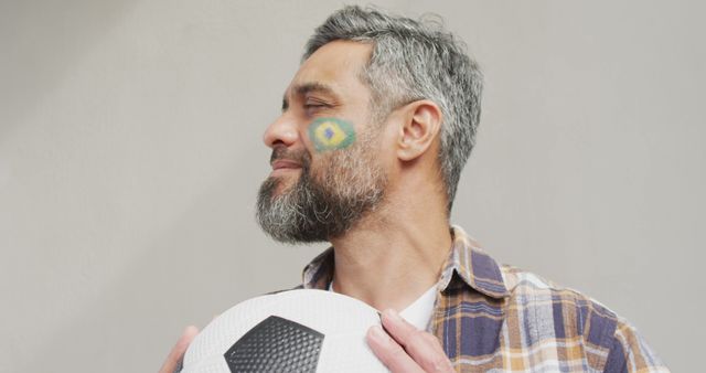 Middle-aged man with a graying beard holding a soccer ball, sporting a casually plaid shirt and having the Brazil flag painted on his cheek while looking to the side with a proud smile. Great for themes involving sports enthusiasm, patriots, fandom, connection to soccer, celebrating national pride, advertising related to sports events or merchandise.