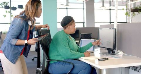 Diverse female coworkers analyzing charts on monitor and taking notes in modern open-plan office