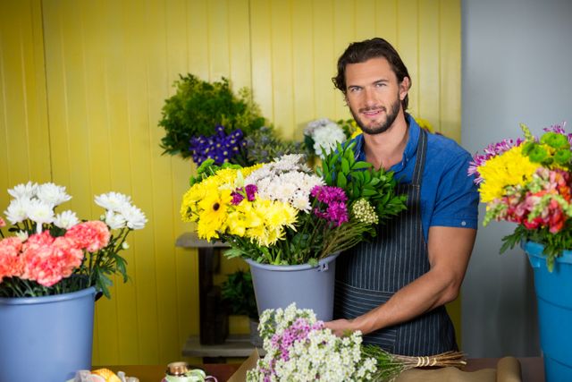 Male florist smiling while holding a bucket of colorful flowers in a flower shop. Ideal for use in articles about small businesses, floral arrangements, customer service, and retail environments. Perfect for advertisements, blogs, and websites related to floristry, gardening, and entrepreneurship.