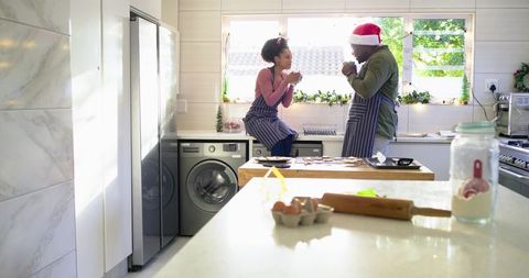 Couple baking cookies in sunlit modern kitchen wearing striped aprons and Santa hat