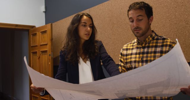 Caucasian couple reviews a blueprint in an office. They are focused on project details, collaborating in a professional environment.