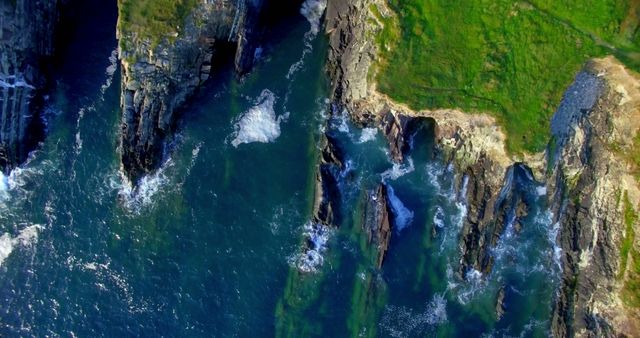 Aerial view captures the dynamic interaction between the ocean waves and rugged cliffs, showcasing nature's raw beauty. The contrast of the lush greenery atop the cliffs against the deep blue sea emphasizes the diverse textures and colors of the coastal landscape.