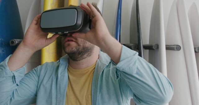 Young man wearing a VR headset, fully immersed in a virtual experience. Background includes several surfboards lined up, indicating a beach setting or surf shop. This image can be used for technology, virtual reality, and surfing-related content. Ideal for promotions or advertisements for VR devices, gaming technology, or surf-related activities. Also suitable for blogs or articles discussing new and innovative ways to experience surfing.