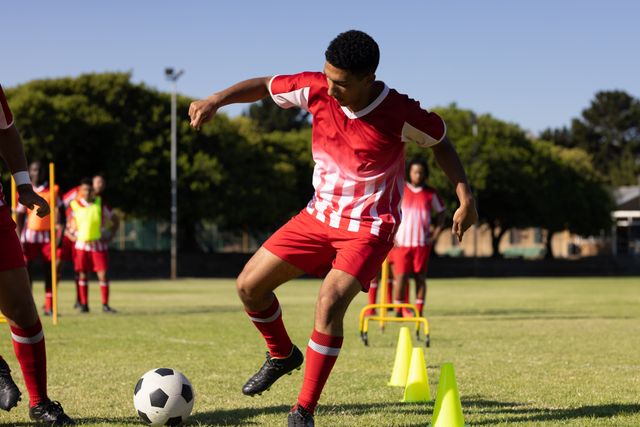 Caucasian player in red uniform dribbling soccer ball around cones on grassy field in trailing drill. Playground, summer, practicing, unaltered, sport, competition, exercising, training and fitness.