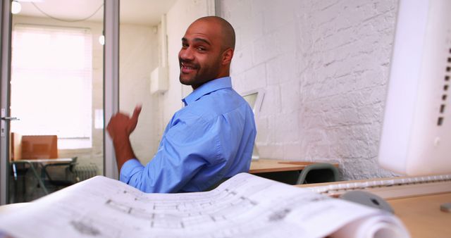 A confident architect in a modern office reviewing a blueprint. He is smiling and looking directly at the camera, creating a friendly and inviting scene. Suitable for use in articles and advertisements related to architecture, design, workspace inspiration, career in architecture, and professional environments.