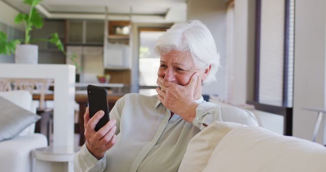 Elderly woman sitting on couch reacting emotionally while looking at smartphone. The environment is a modern, well-lit living room. Suitable for themes related to senior citizens, technology use, family connections, emotional moments, and modern home interiors.