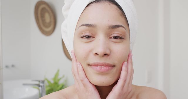 Young woman standing in a bright bathroom with a towel wrapped on her head, gently touching and pampering her face. Suitable for use in skincare, beauty, self-care, and health-related themes and marketing. Can be used for cosmetic product promotions, relaxation, and wellness advertisements and articles on daily skincare routines.