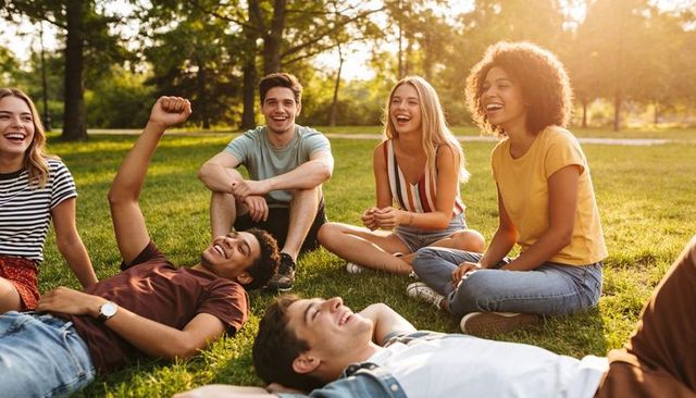 Laughing friends relaxing on park grass at golden hour, sitting in casual summer clothing